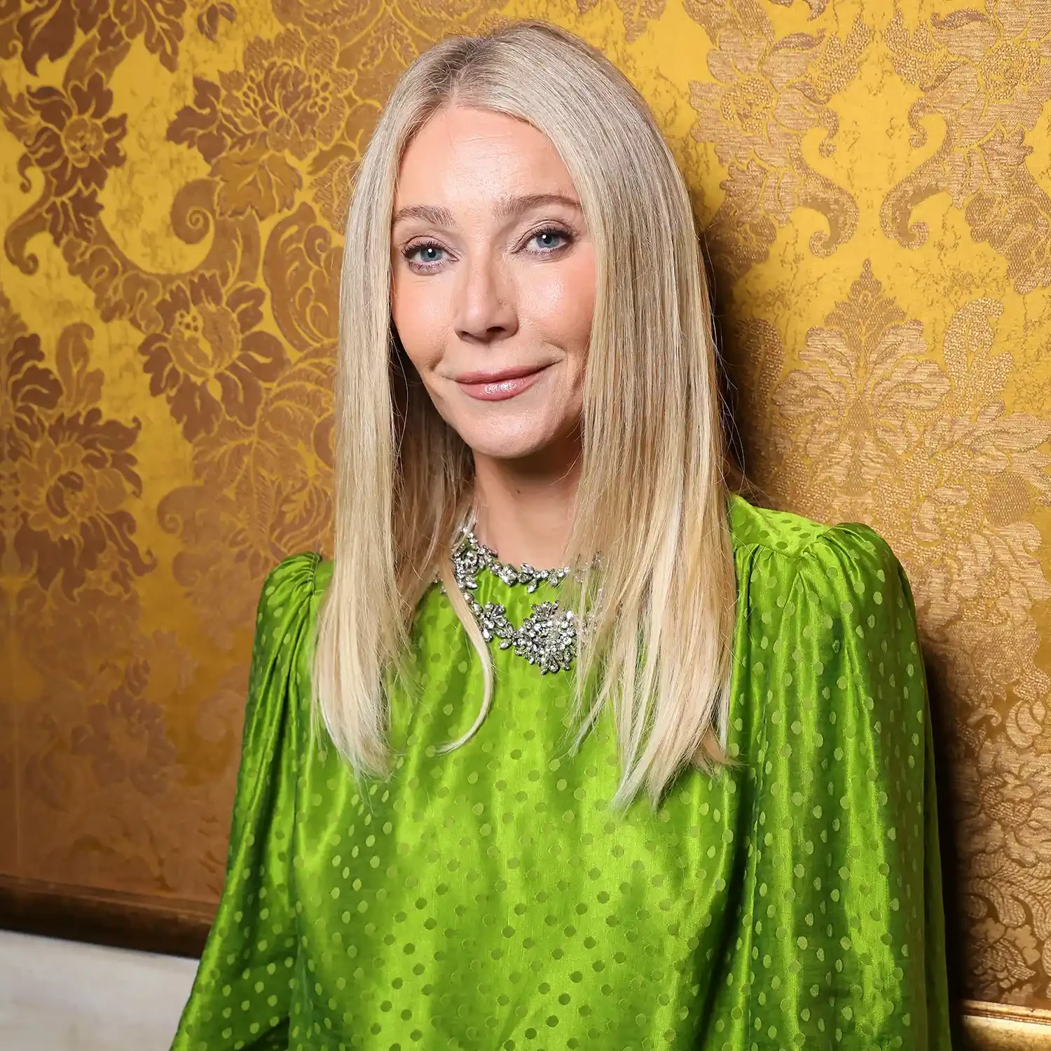 Gwyneth Paltrow diện chiếc váy lụa màu xanh lá nổi bật với bề mặt vải bóng nhẹ và họa tiết chấm nhỏ tinh tế, phần tay phồng nhẹ tạo cảm giác cổ điển xen lẫn hiện đại. Điểm nhấn của trang phục là chiếc vòng cổ kim cương được thiết kế cầu kỳ, mang lại vẻ sang trọng mà không quá phô trương.